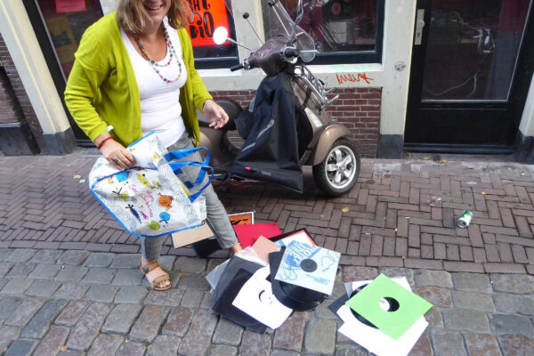 Lachende vrouw met groene cardigan en gekleurde ketting houdt een tas vast waaruit vinylplaten gevallen zijn die op de grond liggen.