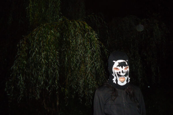 Persoon draagt een zwart masker met een witte schedelprint, staand voor donkere bomen met hangende takken.