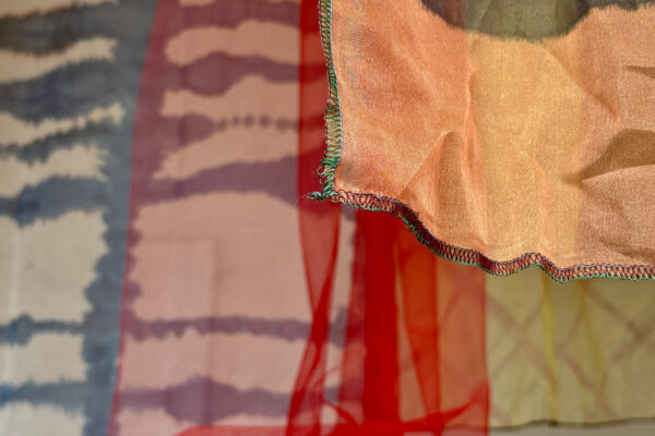 Detail van een gekleurde textielinstallatie met genaaide randen en overlappende transparante lagen in warme tinten.