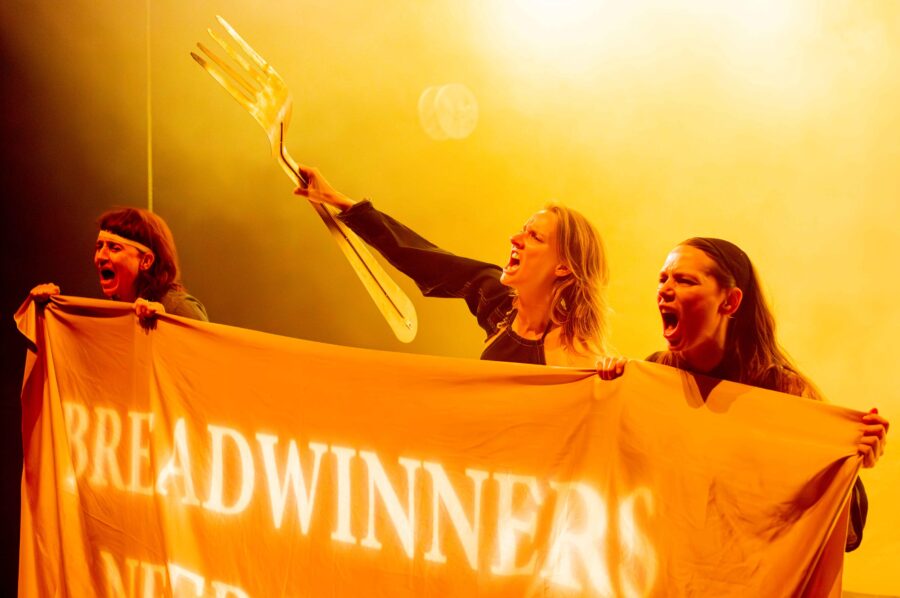 drie vrouwen staan achter een doek waarop het woord 'breadwinners' geprojecteerd is. de vrouw in het midden houdt een groot plastic vork vast, de vrouwen links en rechts houden het doek vast. de drie vrouwen roepen. three women stand behind a piece of cloth on which the word 'breadwinners' is projected. the woman in the middle is holding up a gigantic plastic fork, the women on the left and right hold the piece of cloth. all three women are shouting.