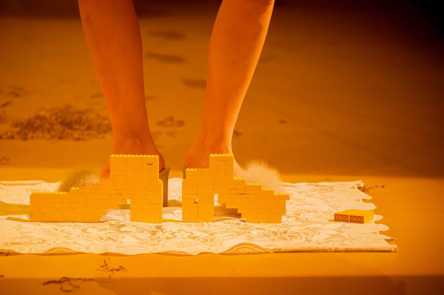 close-up van de onderbenen van een persoon. de voeten zijn verborgen achter twee met legostenen gemaakte schoenen. voeten en schoenen staan op een kanten kleedje. close up of the lower legs of a person. the feet are hidden behind two shoes made of lego. feet and shoes stand on a white piece of lace.