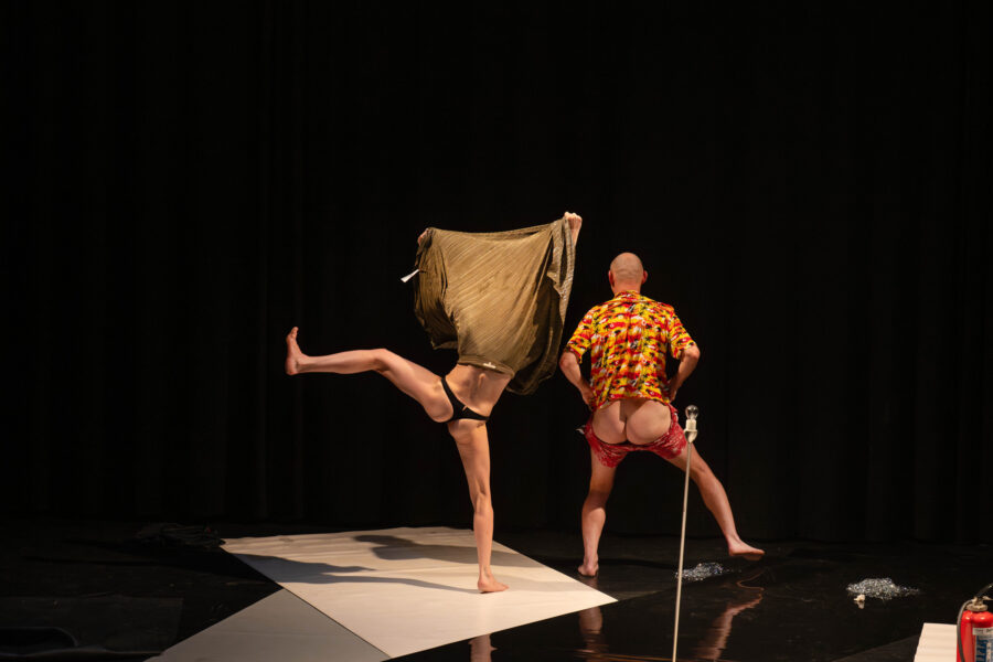Een man en vrouw staan met hun rug naar de camera te dansen, allebei met half ontbloot achterwerk op een zwart theaterpodium. A man and woman dance with their backs to the camera, their asses partly naked, on a black theatre stage.