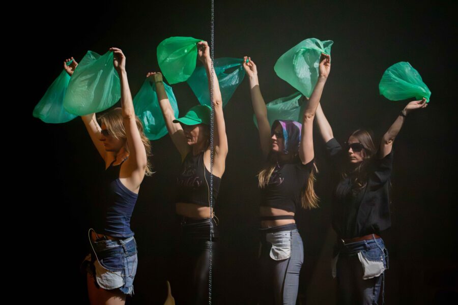 zijwaarts beeld van vier vrouwen die achter elkaar staan en elk twee groene plastic zakken boven hun hoofd houden (een zak in elke hand)sideway image of four women standing behind each other, each holding two plastic bags above their heads (one bag in each hand).